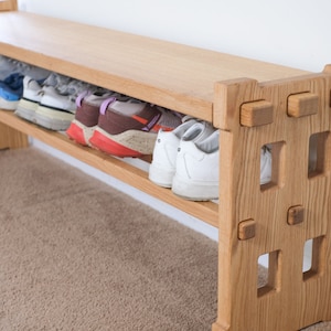 May include: A light-coloured wooden shoe bench featuring a flat top surface and a lower shelf for storage. The side panels have a geometric cutout design with visible joinery details. Several pairs of athletic trainers are stored on the shelf.