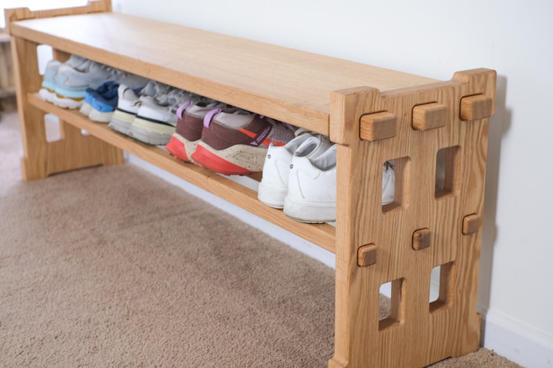 May include: A light-coloured wooden shoe bench featuring a flat top surface and a lower shelf for storage. The side panels have a geometric cutout design with visible joinery details. Several pairs of athletic trainers are stored on the shelf.