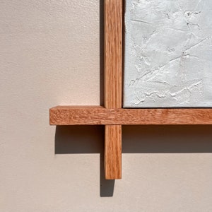 A close-up of a  brown wooden frame corner showcases clean edges around textured white plaster artwork. The minimalist frame contrasts with the organic texture, creating a modern yet natural feel, while light casts subtle shadows on the wall