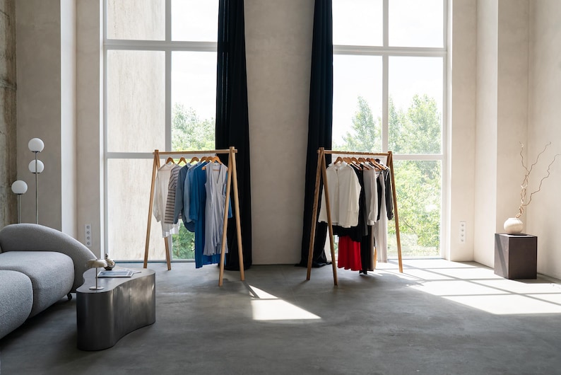 Puede incluir: Dos percheros de madera con ropa colgada. Los percheros est&aacute;n en una habitaci&oacute;n con grandes ventanas y paredes blancas. La ropa es una mezcla de colores y estilos.