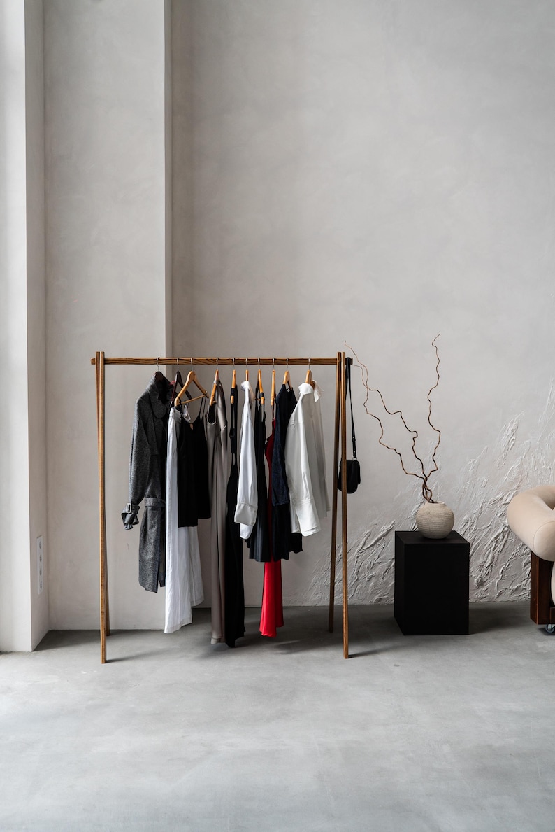 May include: A wooden clothing rack displays a variety of garments, including a long gray coat, dresses, and shirts. The rack is positioned in a minimalist room with a neutral color palette. A small black purse hangs on the side.