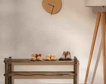 May include: A wooden shoe rack with three shelves, holding pairs of brown and yellow shoes. The rack is in a room with a wooden floor and a white wall with a clock and a floor lamp.
