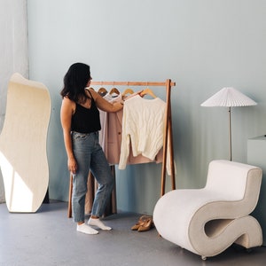 May include: A woman in a black tank top and jeans stands near a wooden clothing rack with a white sweater and a pink blazer. A large, light-colored mirror and a curved white chair are also in the room. The floor is gray.