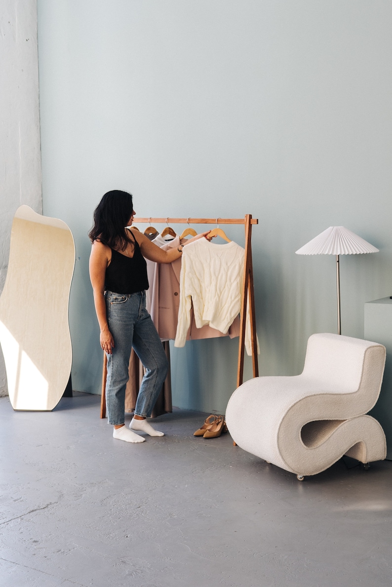 May include: A woman in a black tank top and jeans stands near a wooden clothing rack with a white sweater and a pink blazer. A large, light-colored mirror and a curved white chair are also in the room. The floor is gray.