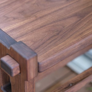 May include: A dark brown wooden bench featuring a smooth rectangular top surface and a sturdy frame constructed with visible joinery. The wood grain is prominent throughout the piece, showcasing natural patterns and deep brown tones in the timber.