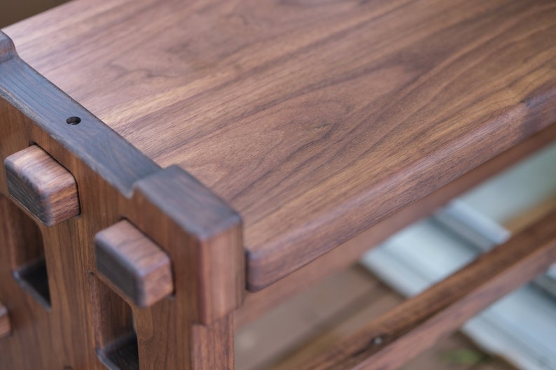 May include: A dark brown wooden bench featuring a smooth rectangular top surface and a sturdy frame constructed with visible joinery. The wood grain is prominent throughout the piece, showcasing natural patterns and deep brown tones in the timber.