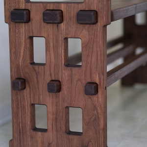 May include: A dark brown wooden bench featuring a rectangular side panel with four vertical cutouts and six protruding square joinery pegs. The bench has a flat top surface and a horizontal support beam connecting the two side panels.