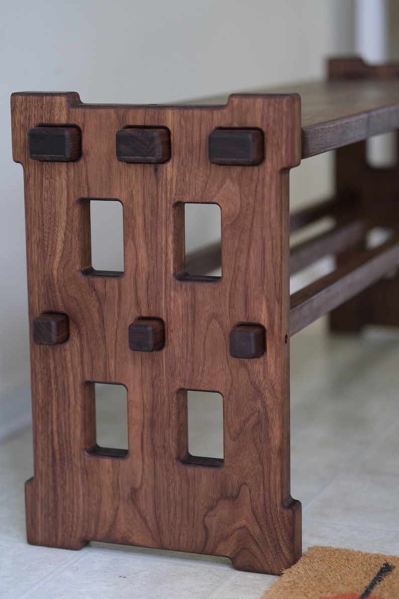 May include: A dark brown wooden bench featuring a rectangular side panel with four vertical cutouts and six protruding square joinery pegs. The bench has a flat top surface and a horizontal support beam connecting the two side panels.