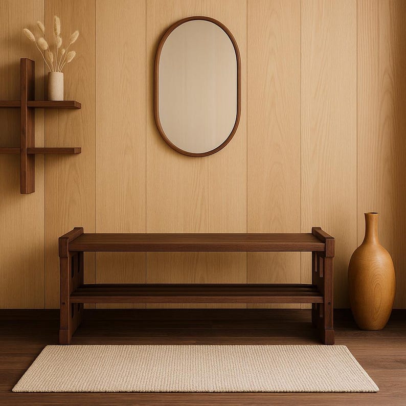 May include: A dark brown wooden bench featuring a slatted lower shelf and a smooth rectangular top. The bench is positioned in front of a light wood panelled wall, accompanied by an oval mirror, a wall-mounted shelf, and a tall wooden vase.