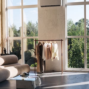 May include: A modern interior with a clothing rack displaying various garments. The rack is brown and holds a long coat, a white shirt, a pink jacket, and a light yellow shirt. A beige sofa and a glass-top table with stone legs are also visible.