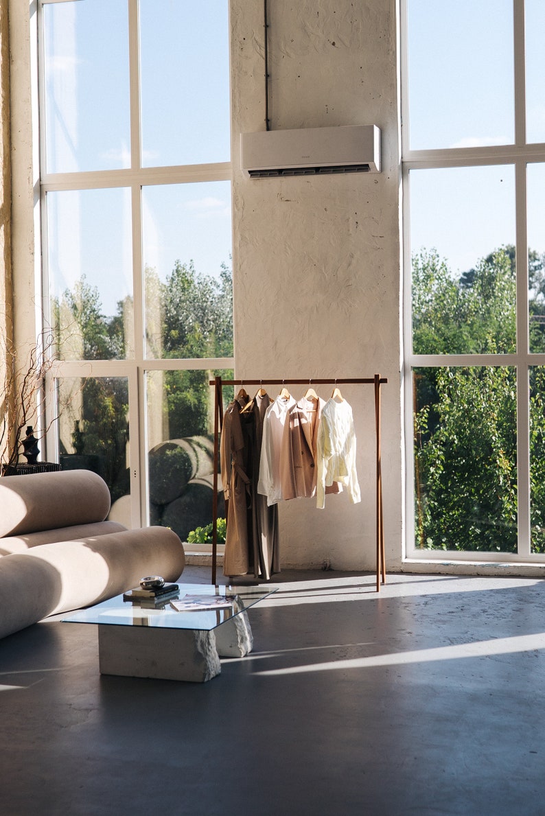 May include: A modern interior with a clothing rack displaying various garments. The rack is brown and holds a long coat, a white shirt, a pink jacket, and a light yellow shirt. A beige sofa and a glass-top table with stone legs are also visible.