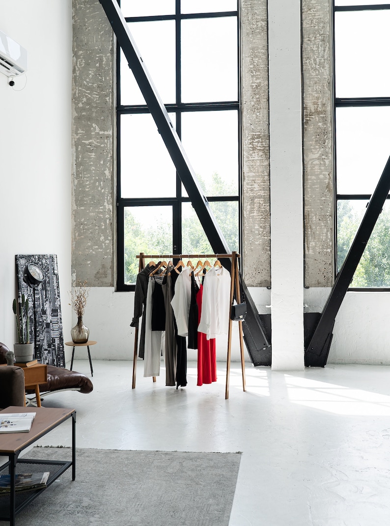 Sturdy ash wood clothing rack, easy to assemble with no tools needed, showcasing chic neutral-toned outfits with a bold red piece. Set in a modern loft space with high industrial windows and minimalist decor.
