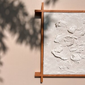 A partially visible brown wooden frame surrounds a white plaster artwork with a textured, organic pattern of raised shapes. The contrast between the smooth frame and the rough artwork is enhanced