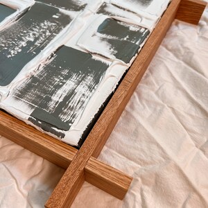 Close-up of textured artwork with gray and white paint patches on a raised surface. Framed in natural wood, the frame extends beyond the edges in a crisscross design, resting on a light fabric background.