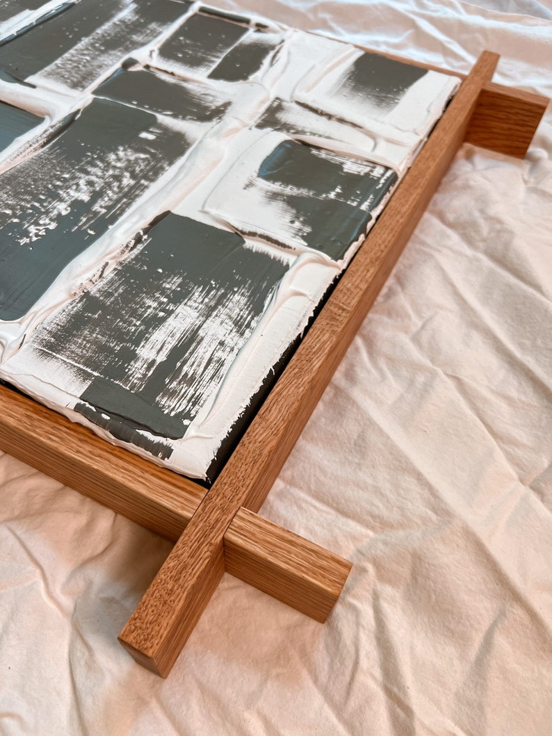 Close-up of textured artwork with gray and white paint patches on a raised surface. Framed in natural wood, the frame extends beyond the edges in a crisscross design, resting on a light fabric background.