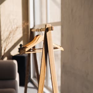 May include: Wooden clothing rack with three wooden hangers. The rack has a minimalist design with an A-frame base and a horizontal bar for hanging clothes. The wood has a natural finish, and the rack is positioned near a window.