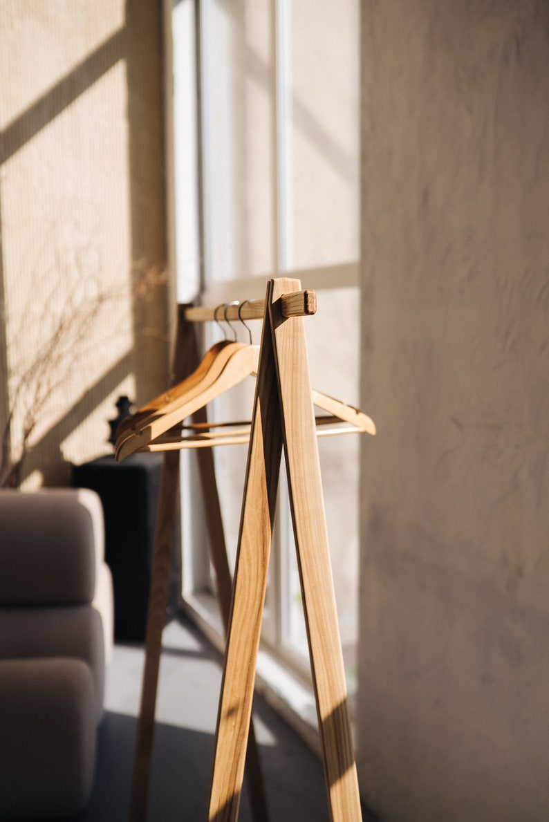 May include: Wooden clothing rack with three wooden hangers. The rack has a minimalist design with an A-frame base and a horizontal bar for hanging clothes. The wood has a natural finish, and the rack is positioned near a window.