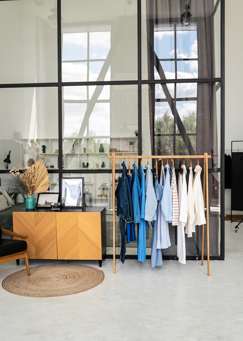 May include: A wooden clothing rack displays a variety of blue and white garments, including jeans, shirts, and a jacket. The rack is in a bright room with a large window and a wooden cabinet with a chevron pattern.