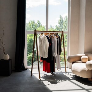 Puede incluir: Un perchero de madera con una variedad de ropa colgada. El perchero est&aacute; en una habitaci&oacute;n con una ventana grande y un sof&aacute; blanco.