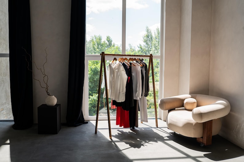 Puede incluir: Un perchero de madera con una variedad de ropa colgada. El perchero est&aacute; en una habitaci&oacute;n con una ventana grande y un sof&aacute; blanco.