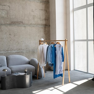 May include: A wooden clothing rack displays a selection of apparel in a modern, minimalist interior. The garments include a white shirt, a striped top, a blue button-down shirt, and a blue dress. The room features a gray sofa, a dark metal coffee table, and a large window.