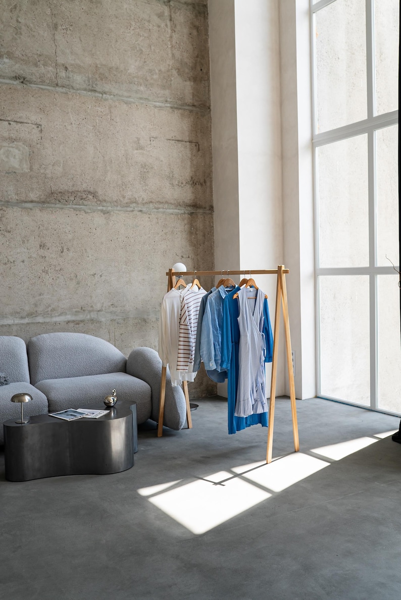 May include: A wooden clothing rack displays a selection of apparel in a modern, minimalist interior. The garments include a white shirt, a striped top, a blue button-down shirt, and a blue dress. The room features a gray sofa, a dark metal coffee table, and a large window.
