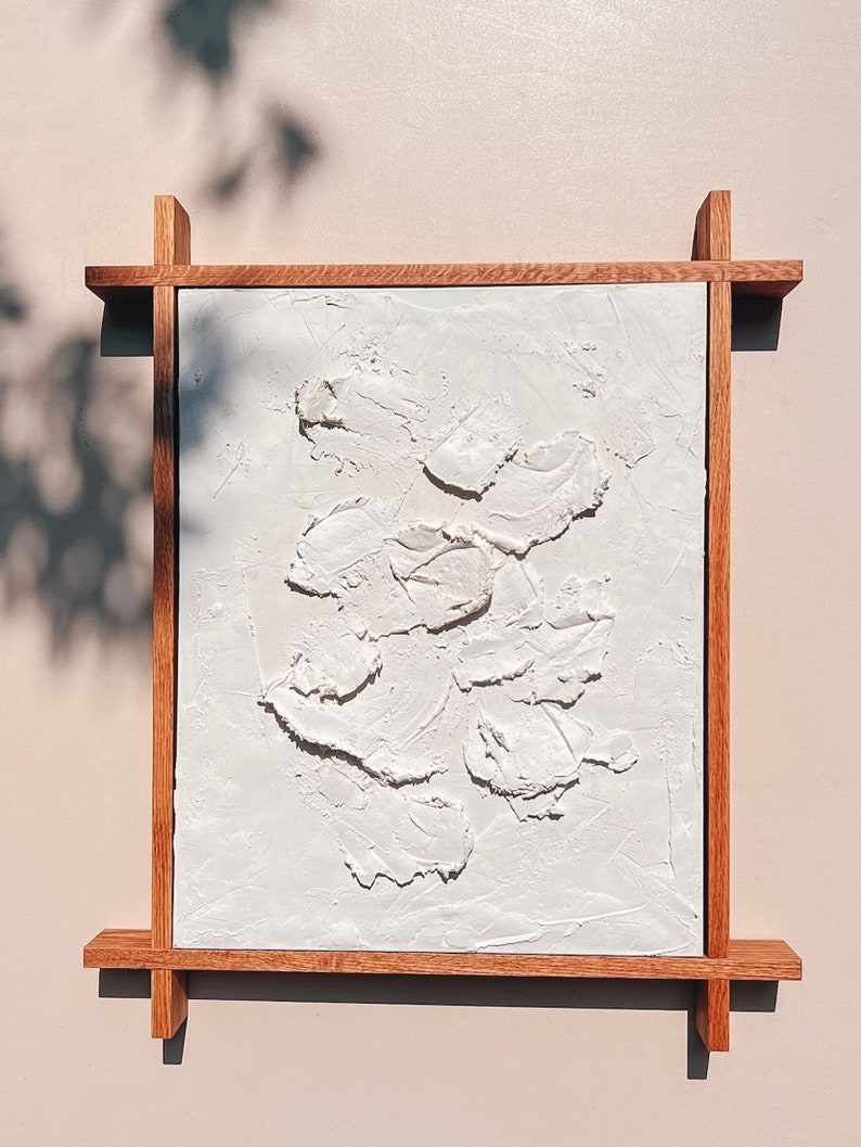 A square wooden frame made from rich brown wood with clean, minimalist lines. Inside is a textured white plaster artwork featuring organic shapes. The rough texture contrasts with the sleek frame, blending modern and rustic styles.