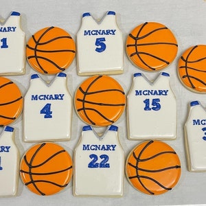 Pode incluir: Uma coleção de biscoitos decorados em forma de camisolas de basquetebol e bolas de basquetebol. As camisolas são brancas com letras e números azuis, e as bolas são laranja com linhas pretas. Os biscoitos estão dispostos numa superfície clara.