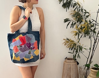 May include: A blue denim tote bag with a patchwork design of colorful fabric scraps. The bag has a long strap and is being held by a person.