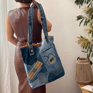 Può includere: Una grande borsa tote in denim blu realizzata con jeans riciclati. La borsa ha un design patchwork con diverse tonalità di denim blu e dettagli in pelle marrone. La borsa ha due lunghe maniglie in denim.