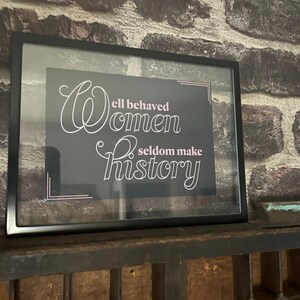 May include: Framed print with the quote "Well behaved women seldom make history" in white script on a dark background. The frame is black, and the print is displayed on a wooden shelf against a brick wall.