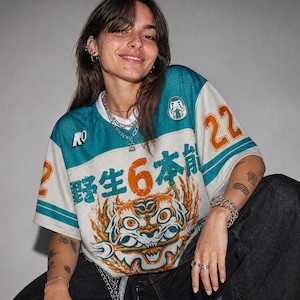 May include: A short-sleeved, mesh-textured jersey in white and teal, featuring the orange number 22 on the sleeves, the text "野生6本能" across the chest, and a graphic of a stylized tiger face surrounded by orange flames on the lower front.