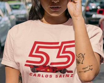 Puede incluir: Una camiseta rosa con un gráfico de coche de carreras rojo y el número 55. El texto "Carlos Sainz" está impreso debajo del coche. La modelo lleva una gorra de béisbol roja con un gráfico de calavera y huesos cruzados blancos.