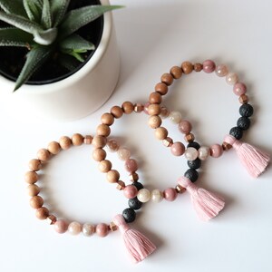 May include: Three bracelets with pink tassels. Each bracelet is made with wooden beads, pink and clear gemstone beads, and black lava rock beads.