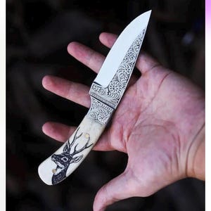 May include: A fixed-blade knife featuring a silver-colored metal bolster with intricate scrollwork engravings. The handle is made of a light-colored material and displays a detailed black ink illustration of a deer head with antlers.
