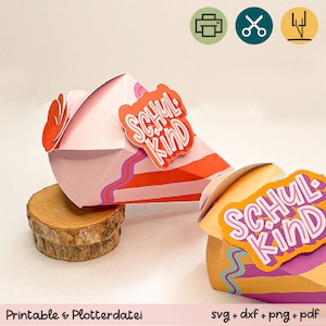 May include: Two colorful, geometric paper gift boxes with the text "SCHUL KIND" in orange and white. The boxes are pink, orange, and yellow with purple accents. The phrase "Printable & Plotterdatei" is at the bottom.