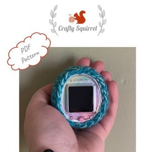 May include: A teal and white crocheted cover for a Tamagotchi, a handheld digital pet. The Tamagotchi is silver and pink with a black screen.