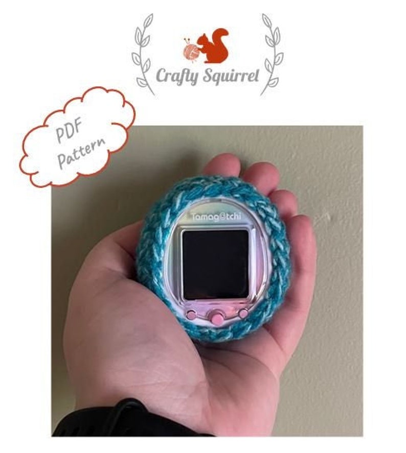 May include: A teal and white crocheted cover for a Tamagotchi, a handheld digital pet. The Tamagotchi is silver and pink with a black screen.