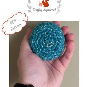 May include: A teal and white crocheted ball in a person's hand. The ball is made with a textured yarn and has a circular shape. The text "PDF Pattern" is visible in the background.