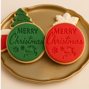 Puede incluir: Una bandeja dorada contiene galletas navideñas festivas. Dos galletas redondas, una verde y otra roja, muestran el texto "MERRY Christmas" y diseños de copos de nieve y renos. Otras galletas incluyen un árbol y un copo de nieve.