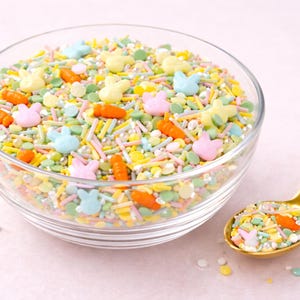 May include: A glass bowl brimming with Easter sprinkles, featuring bunny and carrot shapes. A gold spoon sits alongside, also laden with sprinkles. The sprinkles are in pastel shades, ideal for decorating cakes and biscuits.