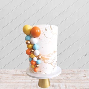 May include: A white cake with gold marbling and a cascade of colorful candy balls in yellow, orange, blue, and white. The cake is on a white cake stand with a gold base.