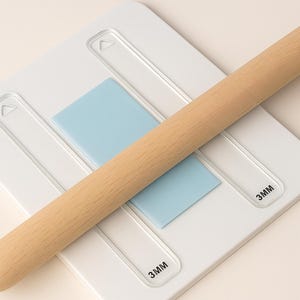 May include: A light-colored wooden rolling pin rests on a white pastry board with a light blue insert. The board has recessed areas and the text "3MM" is printed on the board.