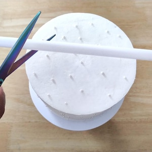 Può includere: Una torta bianca con decorazioni a perle viene tagliata con forbici iridescenti. Un tassello bianco viene tagliato a misura. La torta è su un vassoio bianco, su una superficie di legno marrone chiaro.