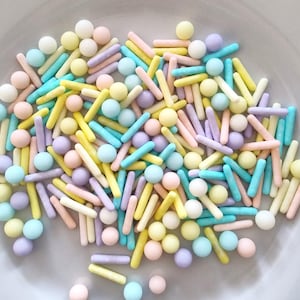 Pode incluir: Sprinkles de cores pastel em uma mistura de formas redondas e longas. Os sprinkles são em tons de azul, amarelo, rosa e roxo.