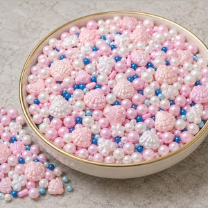 May include: A round white bowl filled with a mix of edible sprinkles including light pink and white pearl-shaped spheres, small blue spheres, white star-shaped pieces, and light pink seashell-shaped pieces, with a small pile of the same mix nearby.