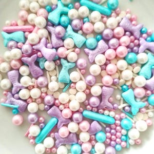 Könnte beinhalten: Eine bunte Mischung aus Streuseln zum Thema Meerjungfrau. Der Mix enthält weiße, rosa, lila und türkisfarbene perlenartige Kügelchen sowie Streusel in Form von Meerjungfrauenschwänzen und Stäbchen. Ideal für Kuchen und Gebäck.