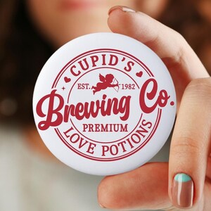 May include: White button with a red circle design. The circle says "Cupid's Brewing Co." with a red heart and a cupid with an arrow inside. It also says "Est. 1982" and "Premium Love Potions".