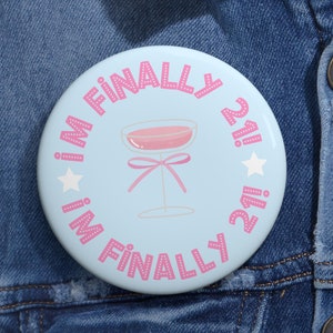 May include: A round pinback button with a light blue background featuring a pink cocktail glass illustration with a bow. The text "I'M FINALLY 21!" is printed in pink dotted lettering around the center, accented by two white stars.