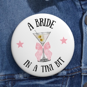 May include: A white button with a pink bow and a martini glass with olives. The text on the button reads "A Bride in a Tini Bit".